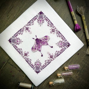 May include: A white fabric with a purple embroidered design of a moth with intricate details. The moth is surrounded by a square frame of purple floral patterns.