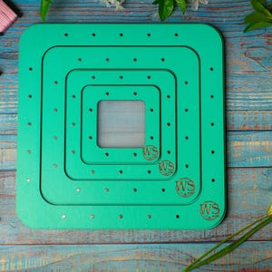 Op de afbeelding: Een teal-kleurige, vierkante houten borduurgaren organizer met meerdere verzonken niveaus. De organizer heeft kleine gaatjes voor het vasthouden van draad en de initialen "WS" zijn in de hoeken gegraveerd. Spoelen garen in verschillende kleuren liggen verspreid.