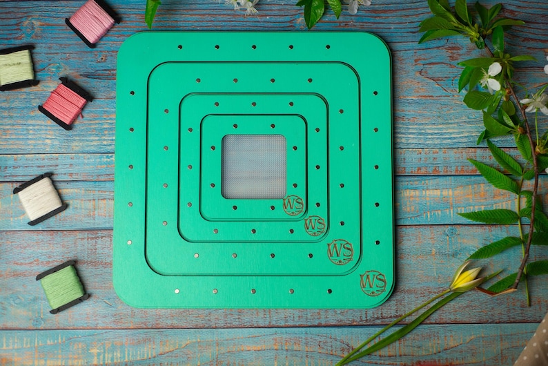 Op de afbeelding: Een teal-kleurige, vierkante houten borduurgaren organizer met meerdere verzonken niveaus. De organizer heeft kleine gaatjes voor het vasthouden van draad en de initialen "WS" zijn in de hoeken gegraveerd. Spoelen garen in verschillende kleuren liggen verspreid.