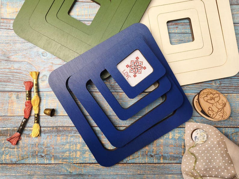Op de afbeelding: Houten borduurringen in groen, blauw en cr&egrave;me, met een kruissteek met het woord "LOVE". Borduurgaren in rood en geel, een vingerhoed en een houten bloemenontwerp zijn ook aanwezig.
