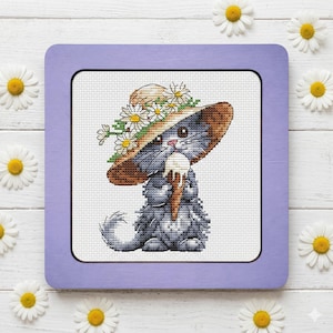 May include: A cross-stitch embroidery featuring a gray kitten wearing a wide-brimmed straw hat adorned with white daisies. The kitten is holding a white ice cream cone. The embroidery is set within a square, light purple wooden frame.