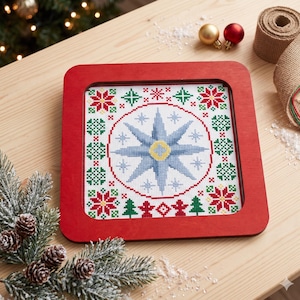 May include: A square, red-framed cross-stitch piece with a central blue starburst design. The piece is surrounded by a border of red flowers, green trees, and snowflake-like patterns. The piece is displayed on a wooden surface with Christmas decorations.