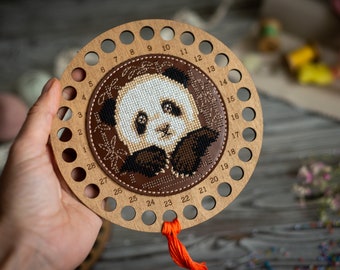 May include: A circular wooden thread organizer featuring a cross-stitched panda design in the center. The light brown wood frame has twenty-eight numbered holes along the perimeter for organizing embroidery floss, with a bright orange thread attached.
