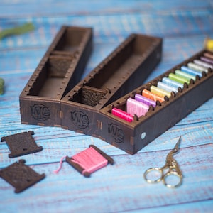 May include: Three wooden thread organizers with lids, each with a different number of compartments. The organizers are filled with colorful embroidery floss. The organizers have the text "WS" engraved on the side.