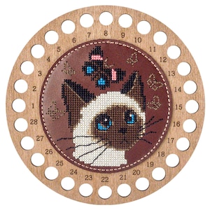 Peut inclure: Un organisateur de fils circulaire en bois avec un empiècement central en cuir marron orné d'un motif au point de croix représentant un chat siamois et un papillon. Le cadre en bois comporte 28 trous numérotés pour organiser le fil.