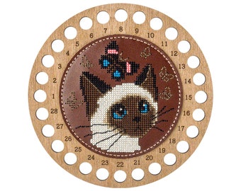 May include: A circular wooden thread organizer featuring a brown leather center patch with a cross-stitch design of a Siamese cat and a butterfly. The wooden frame has 28 numbered holes around the perimeter for organizing embroidery floss.
