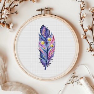 May include: A purple and pink feather with yellow dots and a galaxy design, embroidered on white fabric in a wooden embroidery hoop.