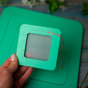 Op de afbeelding: Een groene houten borduurring met een naald en draad, geplaatst op een grotere groene vierkant. De ring wordt door een hand vastgehouden. Andere knutselspullen zijn zichtbaar op de achtergrond.