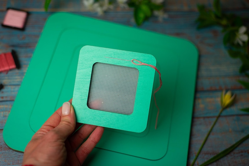 Op de afbeelding: Een groene houten borduurring met een naald en draad, geplaatst op een grotere groene vierkant. De ring wordt door een hand vastgehouden. Andere knutselspullen zijn zichtbaar op de achtergrond.