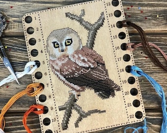 May include: A rectangular wooden thread organizer featuring a cross-stitch owl design. The board has twenty-two numbered holes along the sides for sorting embroidery floss. Strands of white, orange, blue, and brown thread are threaded through the holes.