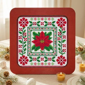 May include: A framed cross-stitch featuring a poinsettia design. The artwork is set within a square, wooden frame. The poinsettia is red with green leaves, surrounded by a border of red and green floral patterns. The frame is a dark reddish-brown color.