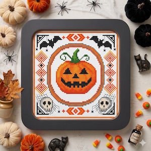 May include: A framed cross-stitch depicting a smiling orange pumpkin with a green stem, surrounded by bats, skulls, and geometric patterns. The Halloween-themed artwork is set against a white background, with decorative pumpkins and spiders.