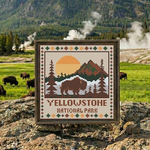 May include: A framed cross-stitch depicting a Yellowstone National Park scene. The design features a bison, mountains, trees, and a setting sun in shades of brown, green, and orange. The words "YELLOWSTONE NATIONAL PARK" are stitched at the bottom.