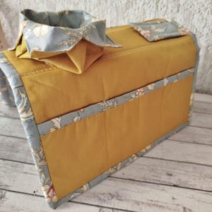 May include: Mustard yellow fabric box with a floral patterned trim and accents. The box has a matching collar and a small square accent piece. The trim and accents feature a light blue background with a floral design.