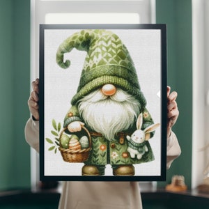 Può includere: Un gnomo verde con la barba bianca tiene un cesto di uova di Pasqua colorate e un coniglietto bianco. Lo gnomo indossa un cappello verde con un motivo bianco e un mantello verde con un motivo floreale.