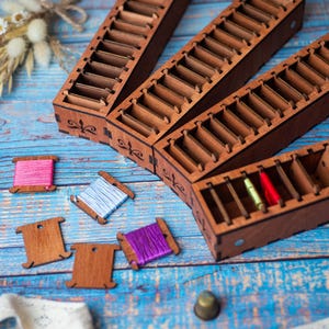 Op de afbeelding: Drie rechthoekige bruine houten organizers met meerdere interne sleuven voor garenopslag. Er zijn verschillende kleine houten spoelen aanwezig, sommige omwikkeld met roze, lichtblauw en paars borduurgaren, naast los groen en rood garen.