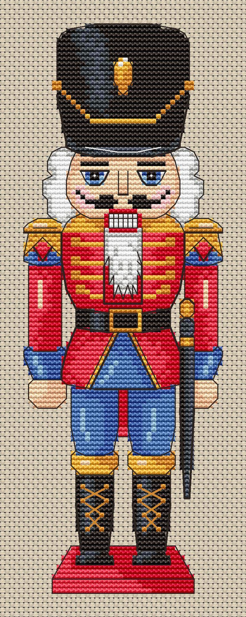 May include: A cross-stitch design of a traditional nutcracker soldier. The figure wears a black hat, red jacket, blue pants, and black boots. The design includes gold, white, and black accents, and a black sword. The background is a neutral beige.