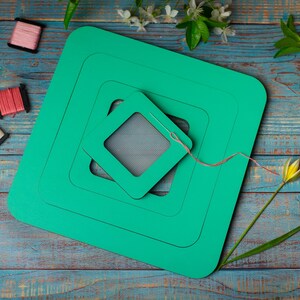 Op de afbeelding: Een turquoise groene knutselmat met een naald en draad, omringd door spoelen garen in verschillende kleuren. De sc&egrave;ne speelt zich af op een verweerd blauw houten oppervlak, met bloemaccenten.