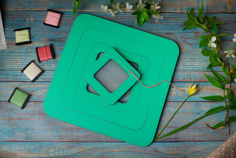 Op de afbeelding: Een turquoise groene knutselmat met een naald en draad, omringd door spoelen garen in verschillende kleuren. De sc&egrave;ne speelt zich af op een verweerd blauw houten oppervlak, met bloemaccenten.