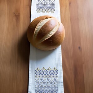 May include: A white table runner with blue and yellow embroidered patterns, featuring a loaf of bread. The runner is rectangular, with vertical stripes and decorative floral and geometric designs. The bread is round and golden brown.