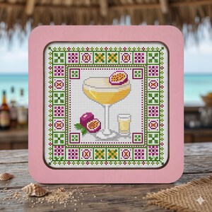 Op de afbeelding: Een vierkant kruissteek borduurwerk met een gele cocktail in een glas op voet, een klein shotglas en passievruchthelften. Het ontwerp is omlijst door een geometrische rand van groene, roze en witte patronen in een lichtroze houten lijst.