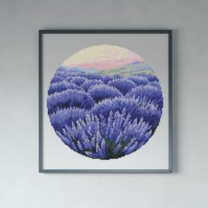 May include: Framed cross-stitch art featuring a circular lavender field under a pastel sky. The artwork is dominated by shades of purple and green, with a light pink and yellow gradient at the top. The frame is a muted gray.