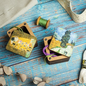 Puede incluir: Dos cajas de madera con tapas con bisagras, cada una con una pintura famosa diferente. Una caja tiene una reproducción de "El Beso" de Gustav Klimt, mientras que la otra tiene una reproducción de "La Noche Estrellada" de Vincent van Gogh. Las cajas están sobre una superficie de madera azul con varios materiales de artesanía.