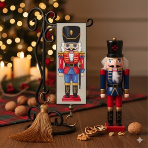 May include: A Christmas-themed image featuring a cross-stitch banner and a wooden nutcracker. The banner depicts a soldier in a red and blue uniform. The wooden nutcracker is in a similar uniform. The background includes a Christmas tree and candles.