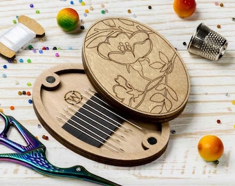 May include: A wooden needle minder with a floral design on the lid. The needle minder is open, revealing a black felt interior with eight needles.