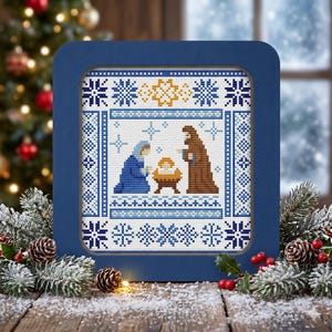 May include: A framed cross-stitch depicting the Nativity scene. The artwork features Mary and Joseph, with the baby Jesus in a manger. The frame is a dark blue, and the cross-stitch has a blue and white snowflake border.