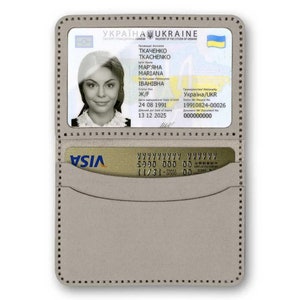 Op de afbeelding: Een grijze leren portemonnee met een transparant plastic venster dat een Oekra&iuml;ens paspoort met de naam "TKACHENKO MAR'YANA IBAHIBHA" en een VISA-creditcard in het onderste vakje toont.