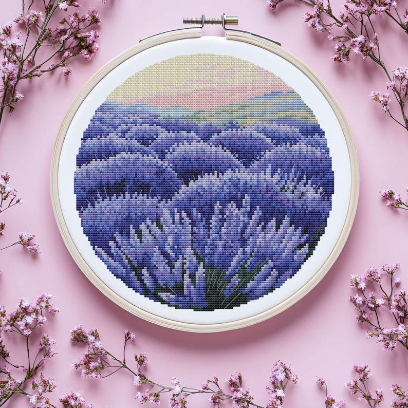 May include: A cross-stitch embroidery of a lavender field in a wooden hoop. The scene features rows of purple lavender under a light pink and yellow sky. The embroidery is set against a pink background with sprigs of pink flowers.