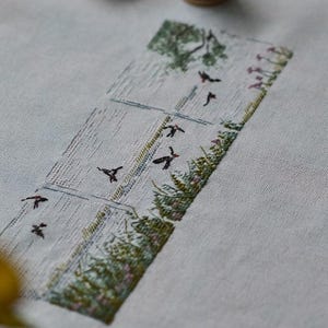 Könnte beinhalten: Gestickte Landschaftsszene mit Feld, Baum und fliegenden Vögeln. Das Kunstwerk ist auf hellblauem Stoff gestickt. Garnrollen im Hintergrund deuten auf ein Handarbeitsprojekt hin.