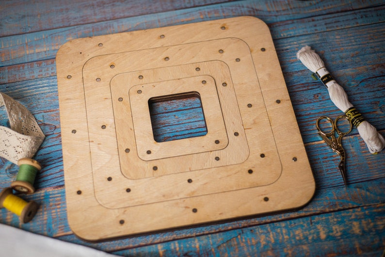 May include: A wooden embroidery hoop with three concentric square frames. The hoop is made of light brown wood and has a natural finish. The hoop is on a blue wooden surface.