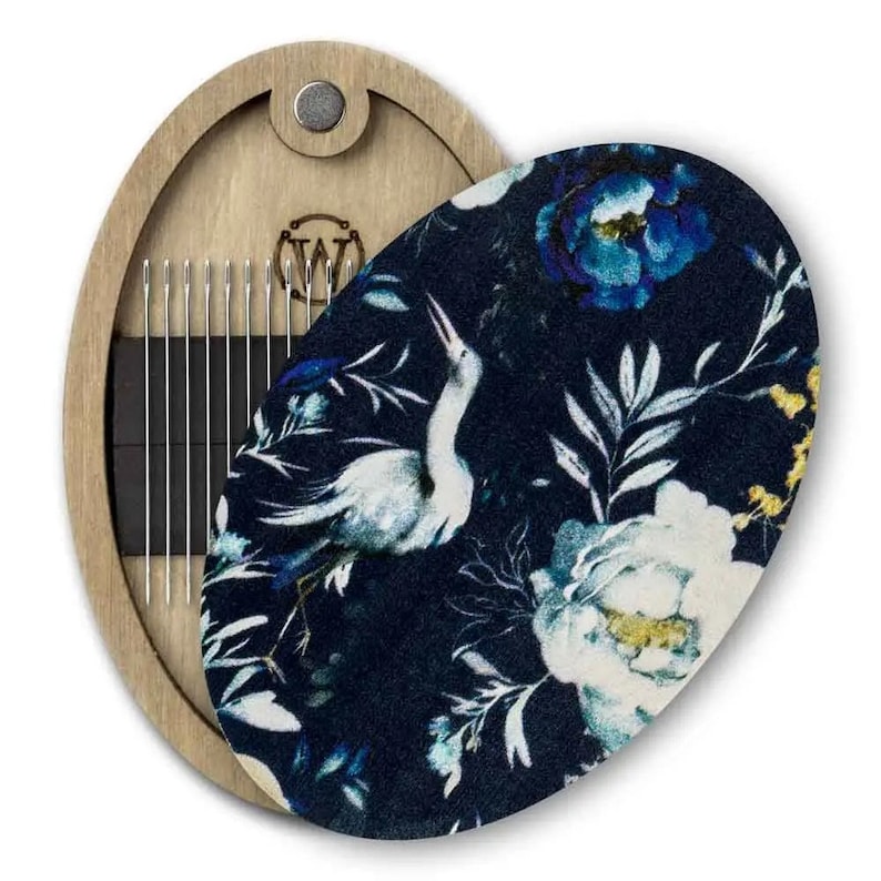 May include: A wooden oval needle case with a magnetic closure. The case has a black felt lining with a floral pattern featuring white birds and blue flowers. The case is open, revealing a row of needles.
