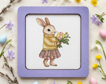 May include: Cross-stitch art of a bunny wearing a yellow sweater and plaid skirt, holding a bouquet of flowers. The artwork is framed in a square, light purple frame. Easter-themed with pastel eggs and flowers.
