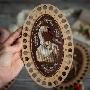 Può includere: Un telaio da ricamo in legno con 32 fori per il filo. Il telaio è decorato con un ovale in pelle marrone che contiene un disegno a punto croce di un cigno bianco con un cigno. Il disegno è circondato da un bordo di piccole perline bianche e cuciture marroni.