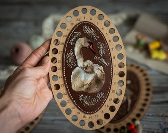 May include: An oval wooden thread organizer featuring a central brown leather patch embroidered with a white swan and cygnet motif. The light wood frame has thirty-two numbered holes along the perimeter for organizing embroidery floss threads.