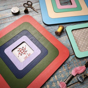 Op de afbeelding: Een verzameling kleurrijke, gelaagde houten frames in rood, groen, blauw en paars. Een kruissteek met het woord "DREAM" is zichtbaar in een van de frames. Borduurgaren, een schaar en een houten spoel zijn ook te zien.
