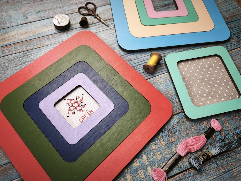 Op de afbeelding: Een verzameling kleurrijke, gelaagde houten frames in rood, groen, blauw en paars. Een kruissteek met het woord "DREAM" is zichtbaar in een van de frames. Borduurgaren, een schaar en een houten spoel zijn ook te zien.