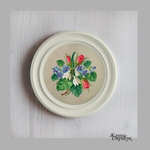 May include: A framed cross-stitch floral design featuring a bouquet of red roses, blue flowers, and green leaves. The artwork is displayed in a round, white frame. The background is a light beige fabric.