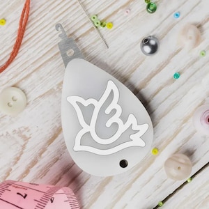 Puede incluir: Un cortahilos gris y blanco en forma de lágrima con un diseño de pájaro. El cortador tiene un gancho de metal y un pequeño agujero. Varios suministros de artesanía, incluyendo cuentas, hilo y una cinta métrica, están esparcidos alrededor.