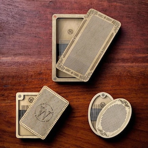 May include: Three wooden boxes with lids, each containing a small needlepoint canvas and needles. The boxes are decorated with floral designs and the lids have a grid pattern. The boxes are on a wooden surface.