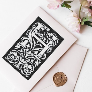 Könnte beinhalten: Eine weiße Karte mit einer schwarz-weißen Illustration des Buchstabens H, umgeben von verzierten floralen Designs. Die Karte liegt auf einem rosafarbenen Umschlag mit einem goldenen Siegel aus Wachs.