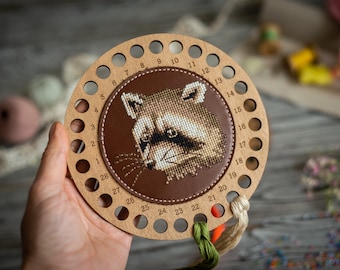May include: A wooden thread organizer with 28 holes numbered 1-28. The organizer has a brown leather center with a cross-stitch image of a raccoon's face.