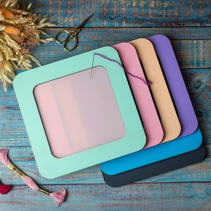 May include: A set of five colorful embroidery hoops with a light blue, pink, peach, purple, and black color scheme. The hoops are stacked on top of each other, with the largest hoop on the bottom and the smallest hoop on top. The hoops are made of wood and have a smooth finish. A needle and thread are visible in the top hoop.