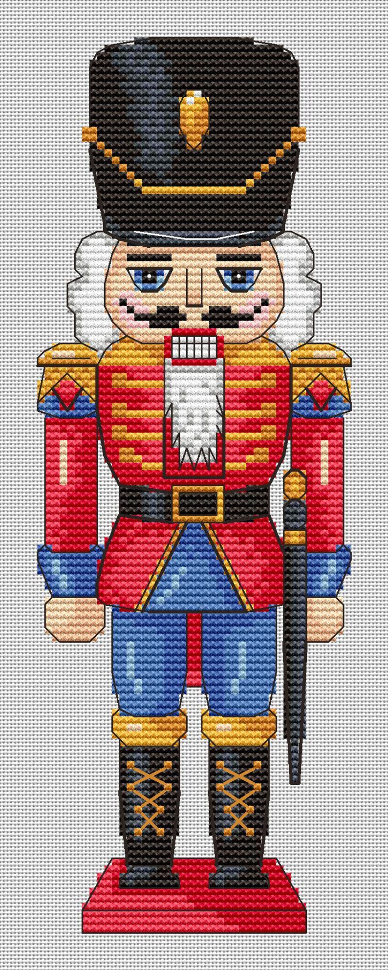 May include: A cross-stitch design of a traditional nutcracker soldier. The figure is dressed in a red jacket with gold accents, blue pants, and a black hat. It holds a black sword and has a white beard and mustache.