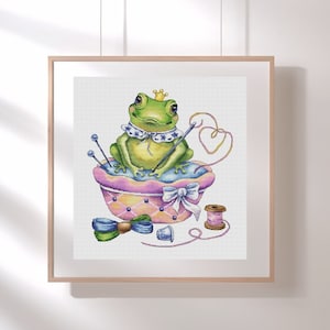Könnte beinhalten: Gerahmtes Kreuzstich-Kunstwerk mit einem grünen Frosch, der eine Krone und einen dekorativen Kragen trägt. Der Frosch sitzt auf einem rosa-gelben Nadelkissen mit Nähnadeln, Faden und einer Schleife. Ein Fingerhut und eine Garnrolle sind ebenfalls zu sehen.