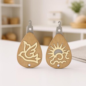 May include: Two wooden teardrop-shaped keyrings. One features a gold-coloured dove design, the other a sun and cloud design. Both have a silver metal top with a hook. The keyrings are made of light brown wood.