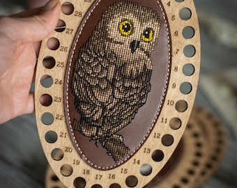 May include: An oval wooden thread organizer featuring a central brown leather inlay with a cross-stitched owl design. The light-colored wood frame has thirty-two numbered holes along the perimeter for organizing embroidery floss and thread.